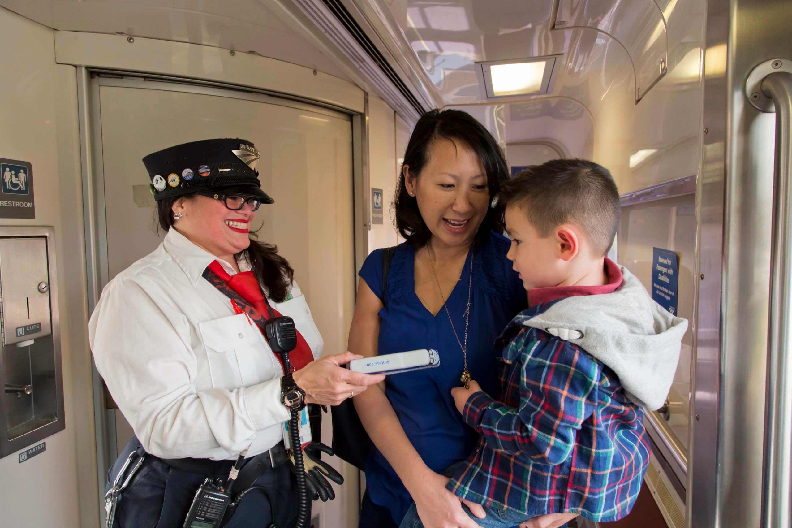 A cheerful Pacific Surfliner employee hands a model train to a young boy riding with his mom.