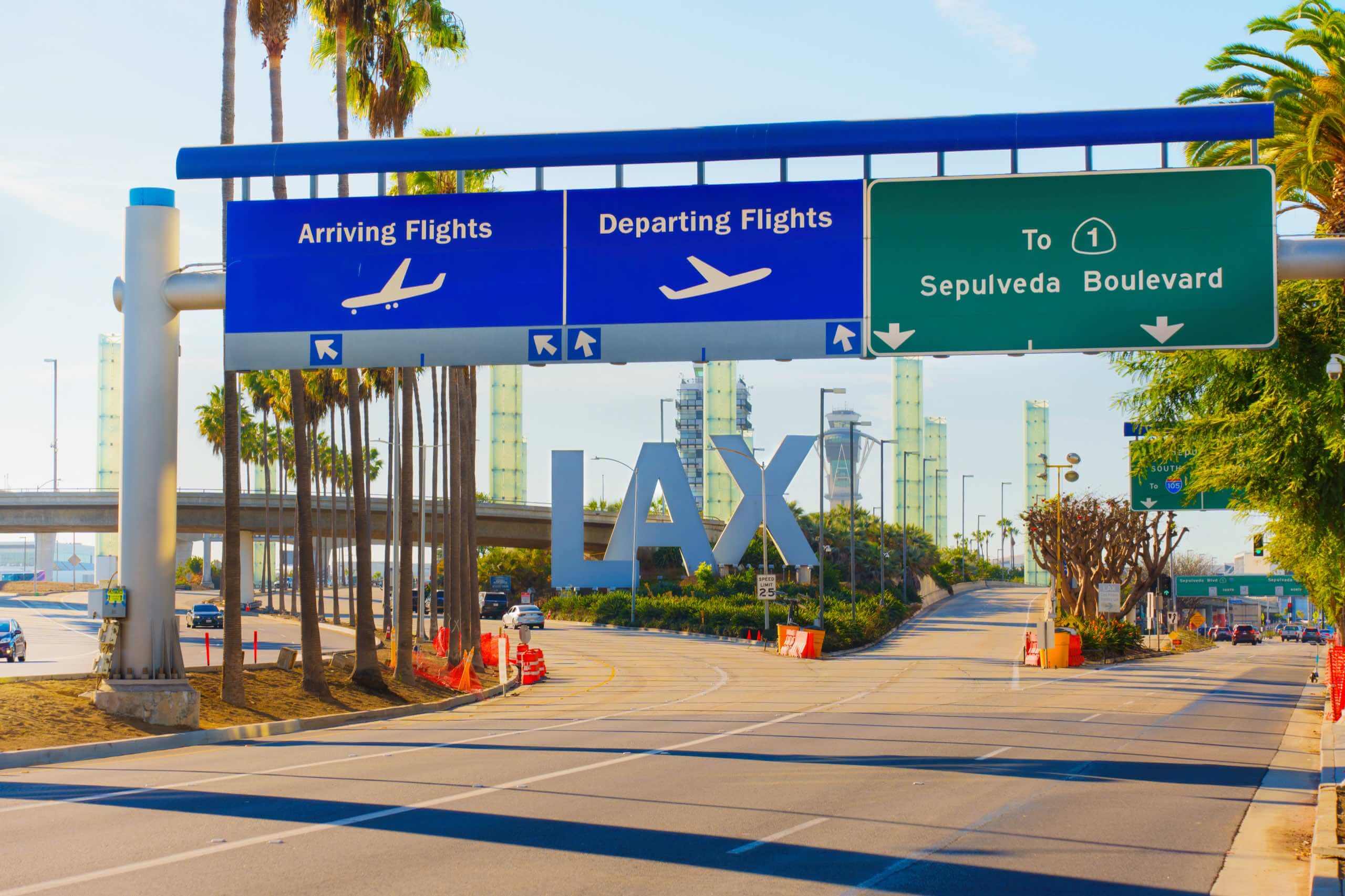 Roadway entrance to Los Angeles International Airport (LAX) with large blue signs for Arriving Flights and Departing Flights, palm trees lining the road, and the iconic LAX letters in the background.